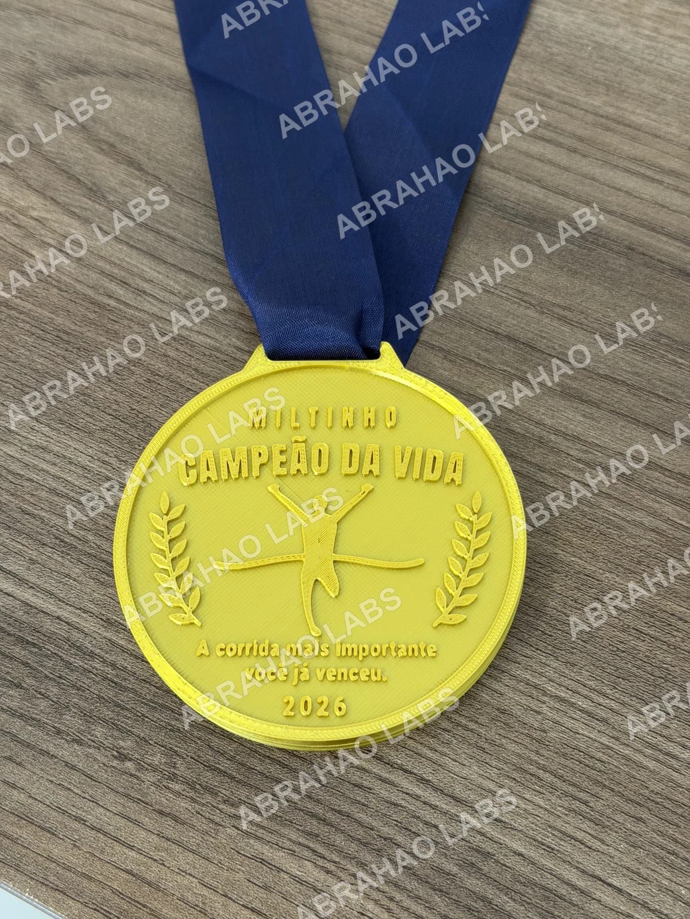 Medalha de homenagem impressa em 3D dourada com nome Miltinho, título Campeão da Vida, corredor e ramos de louro em relevo, fita azul